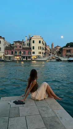 a woman is sitting on the edge of a pier looking at the water and buildings