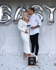 a man and woman standing next to each other in front of balloons that spell out baby