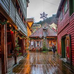 an alley way with wooden buildings and christmas lights on the roof, surrounded by greenery