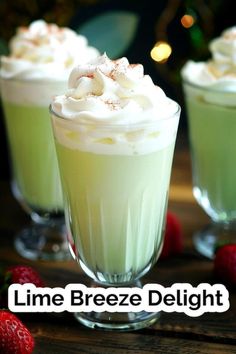 two glasses filled with green liquid and topped with whipped cream next to strawberries on a wooden table