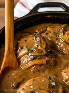 a skillet filled with meat and mushrooms covered in gravy next to a wooden spoon