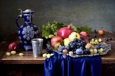 a still life with grapes, apples, and other fruit on a table next to a blue vase