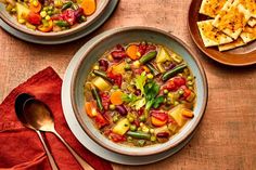 two bowls filled with vegetable soup next to crackers and spoons on a wooden table