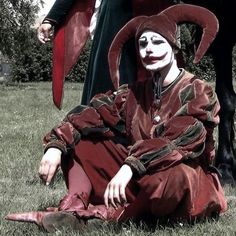 a man dressed as a jester sitting on the ground next to a black horse