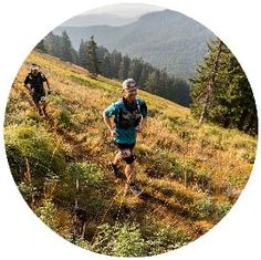 two people are running on a trail in the mountains with trees and grass around them