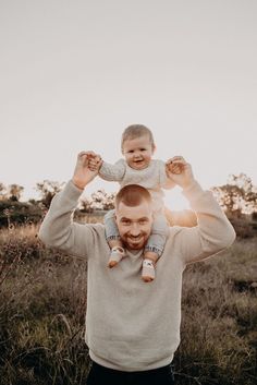 a man holding a baby up in the air with his hands above his head while standing in a field