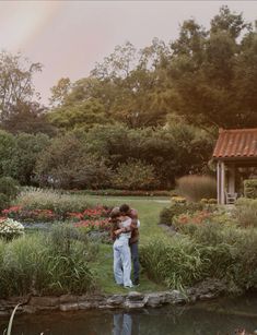 two people embracing each other in the middle of a garden with flowers and trees behind them