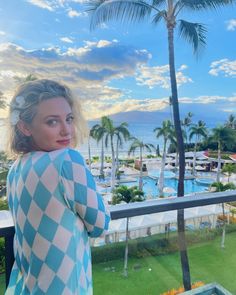 a woman standing on top of a balcony next to a lush green field and palm trees