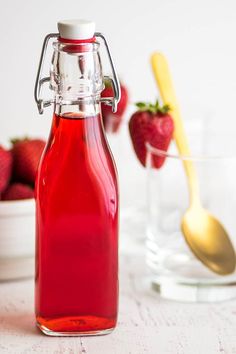 a glass bottle filled with red liquid next to strawberries and a spoon on a table