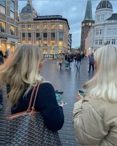 two women looking at their cell phones while walking down the street in front of some buildings