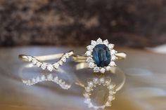 two engagement rings with an oval blue sapphire surrounded by small white diamonds on a reflective surface