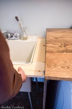 a person standing in front of a sink under a wooden table with utensils on it