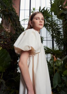 a woman standing in front of plants wearing a white dress with ruffles on the shoulders