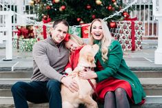 a man and woman sitting next to a dog in front of a christmas tree