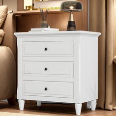 a white night stand with three drawers in front of a couch and lamp on the end table