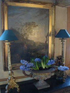 a vase with blue flowers sitting on top of a table next to two lamps and a painting