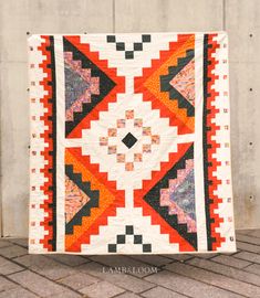 an orange and black quilt sitting on top of a brick floor next to a wall