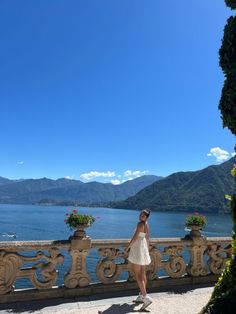 a woman standing on the edge of a balcony next to a body of water with mountains in the background