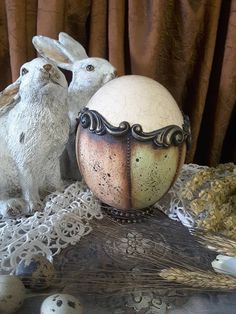 two white rabbits sitting next to an egg on a lace tablecloth with eggs in the background