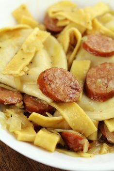 a white bowl filled with pasta and sausage on top of a wooden table next to a fork