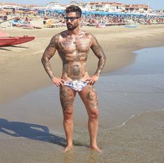 a tattooed man standing in the water at the beach with his hands on his hips