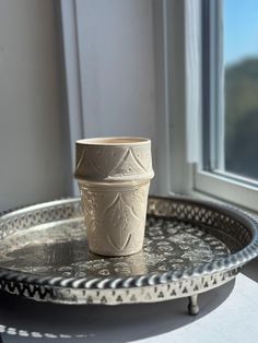 a white cup sitting on top of a metal tray next to a window sill