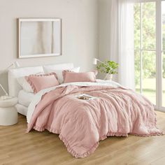 a bed with pink comforter and pillows in a white room next to a window