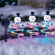 three snowmen with top hats and scarfs on in the snow next to a solar powered lawn light