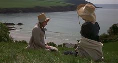 two women sitting on the grass with hats over their heads looking out at the water