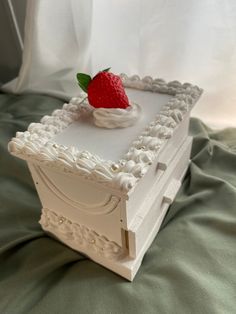 a small white box with a strawberry on top sitting on a green cloth covered bed