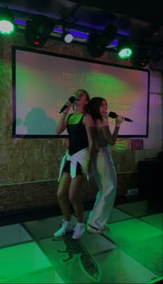 two young women singing into microphones in front of a screen with green lights on it