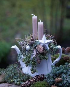 a white tea pot with candles and greenery in it sitting on a table next to pine cones