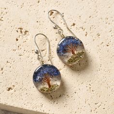 two earrings with trees on them sitting on top of a stone slab next to sand