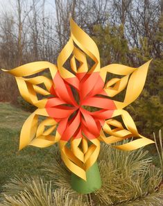 a red and yellow paper flower sitting on top of a grass covered field next to trees