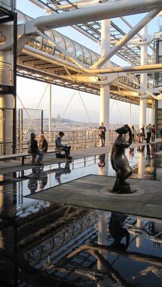 people are walking around in an open area with glass walls and metal structures on the roof