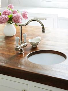 pink flowers in a white vase sitting on top of a wooden counter next to a sink