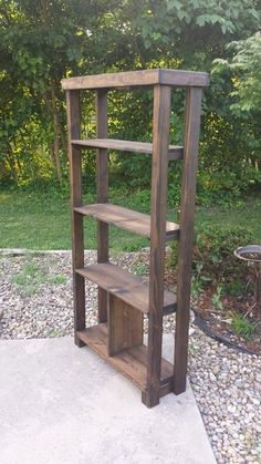 a wooden book shelf sitting on top of a gravel covered ground next to a forest