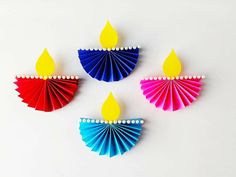 three brightly colored paper diyas on a white surface with one lit candle in the middle