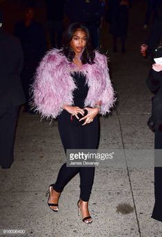 a woman wearing a pink feather jacket and black pants is seen walking down the street