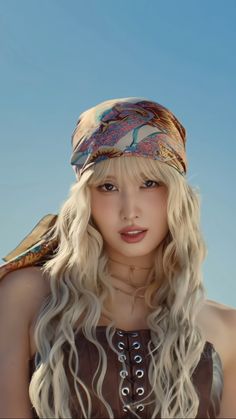 a woman with long blonde hair wearing a bandana and looking at the camera while standing in front of a blue sky