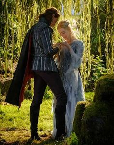 a man and woman dressed in medieval clothing standing next to each other under a willow tree