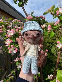 a hand holding a small crocheted doll in front of some trees and flowers