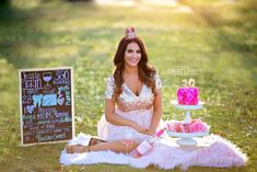 a woman sitting on the grass with a pink cake in front of her and a chalkboard sign behind her