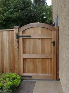 a wooden gate in the side of a building next to a sidewalk and bushes with trees behind it