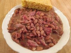 a white plate topped with beans and corn next to a piece of cornbread on top of a wooden table