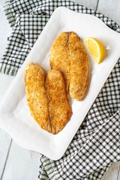 two pieces of fried chicken on a white plate with lemon wedges next to it