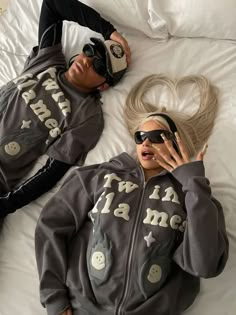 two people laying on top of a bed wearing matching sweatshirts and headphones, one holding a cell phone to her ear