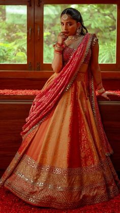 a woman in an orange and gold lehenga standing by a window with her hand on her chin