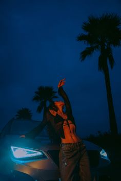 a woman leaning on the hood of a car at night with palm trees in the background