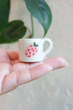 a hand holding a tiny cup with a strawberry painted on the side and green leaves in the background